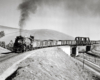 Black-and-white view of steam locomotive with freight train crossing bridge over another railroad and a highway.