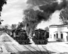 Black-and-white view of steam locomotives heading two narrow-gauge trains on lines converging in a small town