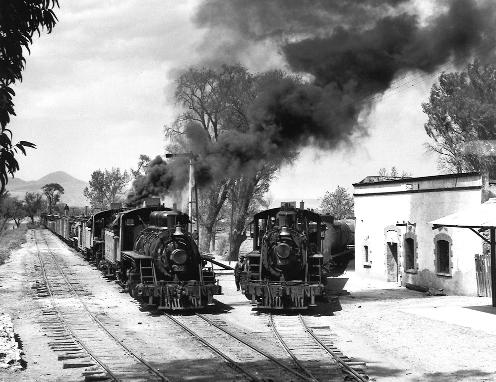 Black-and-white view of steam locomotives heading two narrow-gauge trains on lines converging in a small town