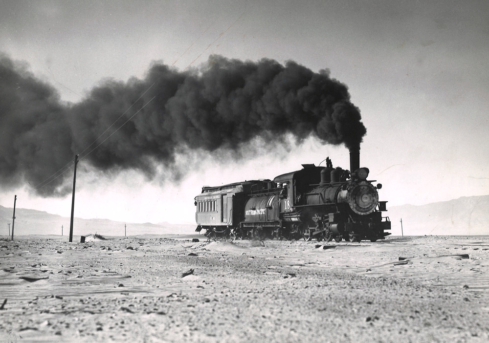 Black-and-white view of 4-6-0 steam locomotive making smoke with 1-car train in desert
