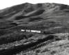 Black-and-white view of road-switcher diesel locomotive with short freight train amid barren hills