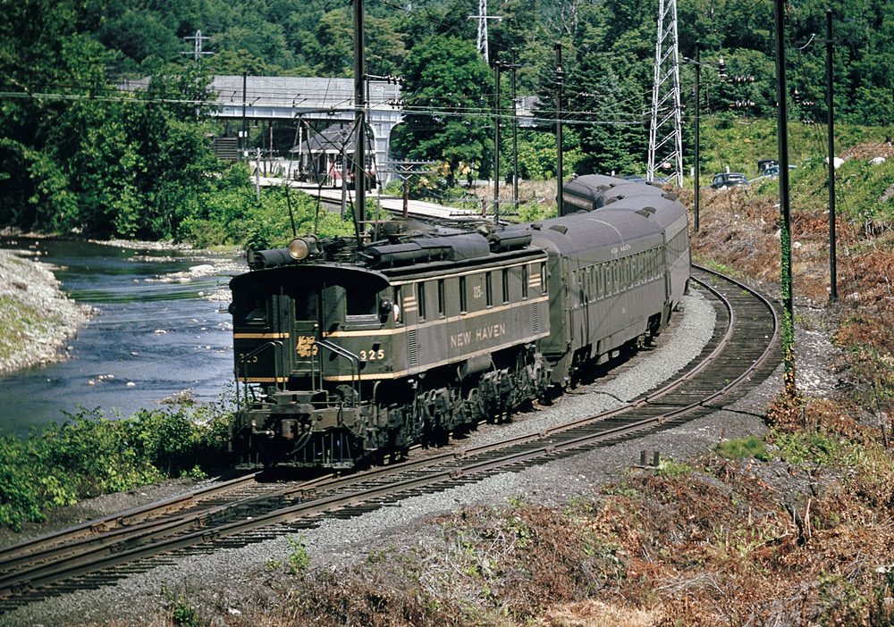 Color photo of box-cab electric locomotive with passenger train on curve beside river