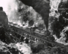 Black-and-white overhead view of 2-6-6-2 steam locomotive with freight train crossing bridge in mountains