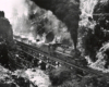 Black-and-white overhead view of 2-6-6-2 steam locomotive with freight train crossing bridge in mountains