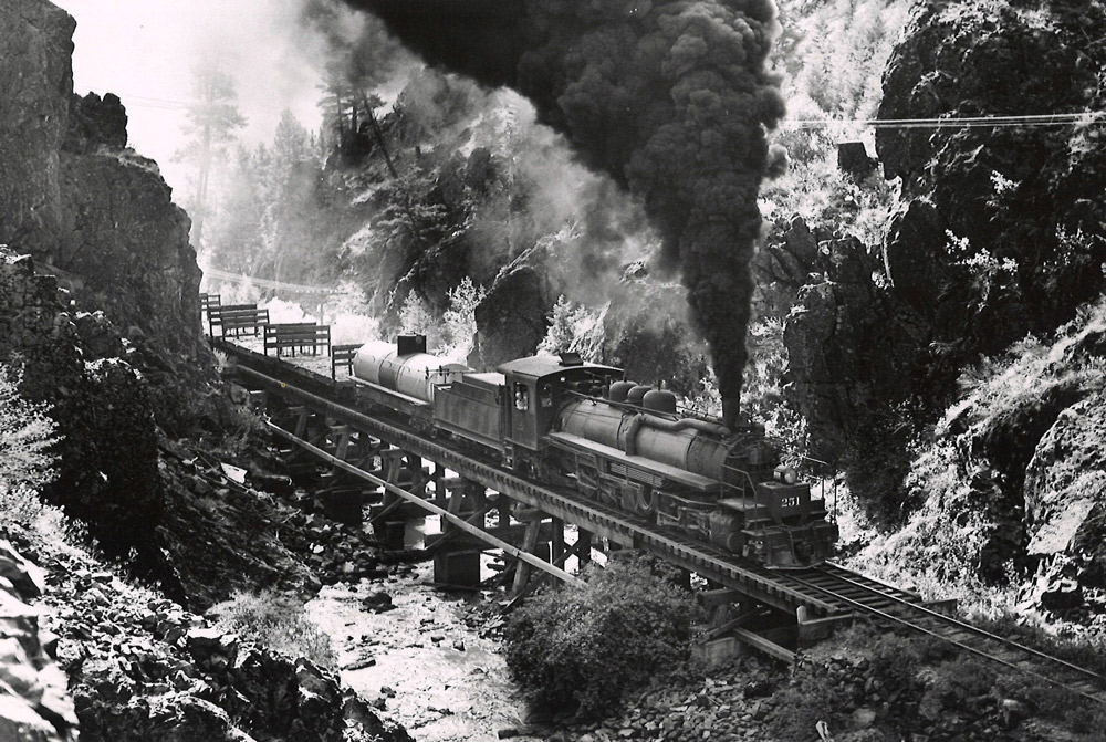 Black-and-white overhead view of 2-6-6-2 steam locomotive with freight train crossing bridge in mountains