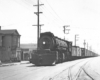 Black-and-white view of steam locomotive with freight train on track in a city street