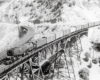 Black-and-white overhead view of steam locomotive with freight train on trestle in mountains