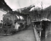 Black-and-white view of steam locomotive with train on dock beside ship