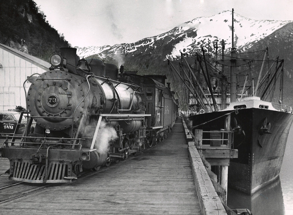 Black-and-white view of steam locomotive with train on dock beside ship