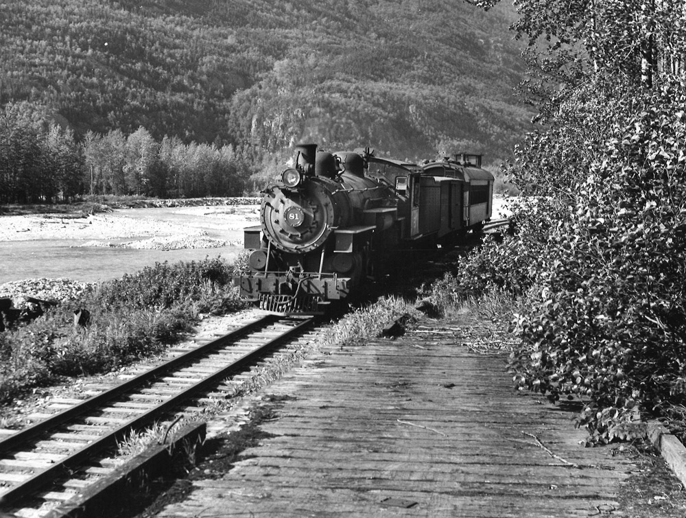 Black-and-white view of 2-8-2 steam locomotive with 2-car train beside river