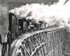 Black-and-white view from behind of two steam locomotives pushing a rotary snowplow on a trestle