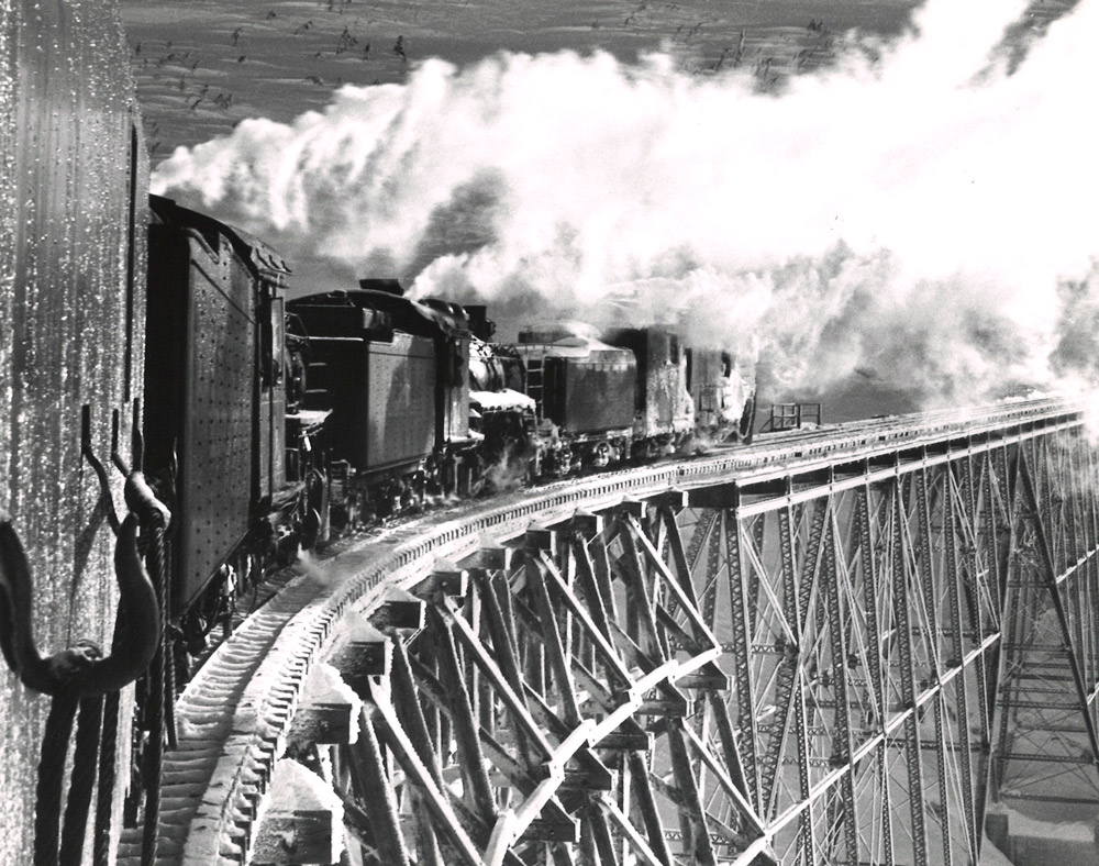 Black-and-white view from behind of two steam locomotives pushing a rotary snowplow on a trestle