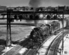 Steam locomotive with passenger train paused underneath a signal bridge in a black and white image.