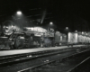 A steam locomotive and passenger train idle under platform lights next to a passenger platform at night.