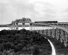 Black and white image of a passenger train crossing a trestle and bridge over a river and made small by the size of them all.