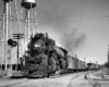 A large steam locomotive leads a freight train beside water towers under clear skies.