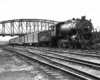 An older steam locomotive leads an express train underneath an arched girder bridge.