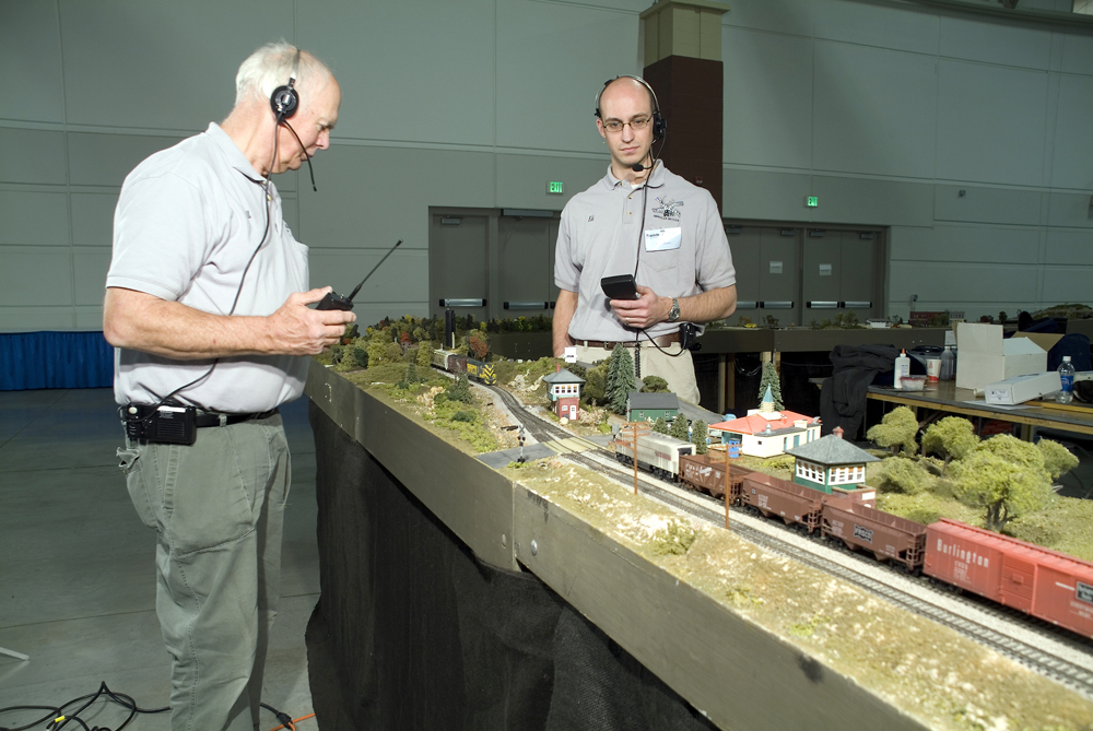 Two men holding throttles operate trains on a modular layout in a convention hall