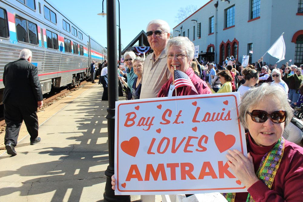 Crowd at train platform including woman holding sign, "Bay St. Louis loves Amtrak"