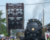 A crowd of people get close to a large steam locomotive that is stopped near an old truss bridge.