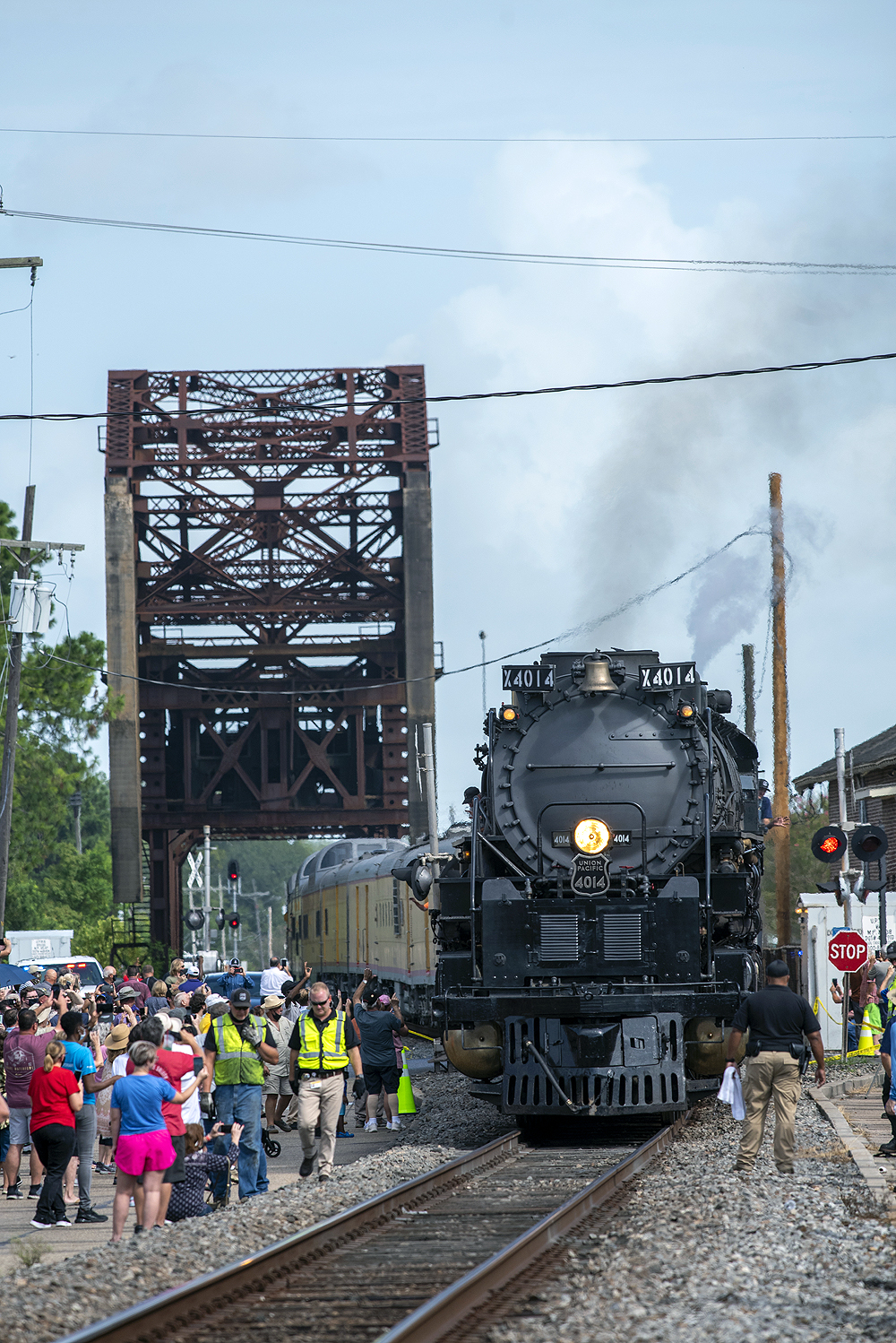 A crowd of people get close to a large steam locomotive that is stopped near an old truss bridge.