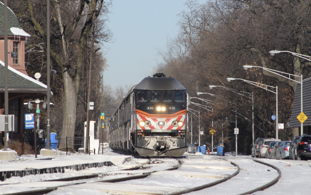 Commuter train led by locomotive with black, orange, and white nose arrives at station on sunny day with snow on the ground.