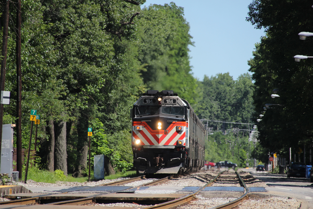 Train arrives at station after traveling down straight lined by trees.