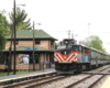 As seen from across tracks, a commuter train arrives at a wood two-story station.