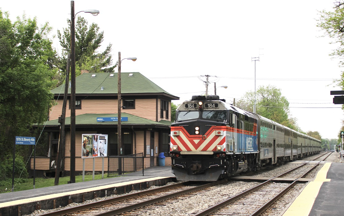 As seen from across tracks, a commuter train arrives at a wood two-story station.