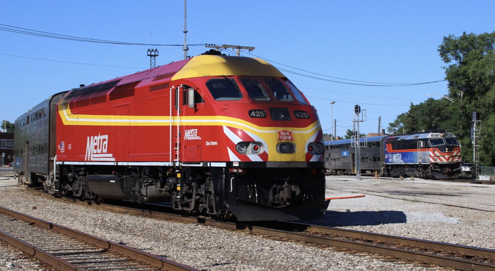 Train with red and yellow locomotive in foreground; train with blue, black, white, and silver locomotive in background.