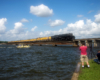 A steam locomotive hauling a yellow passenger train near a lake.