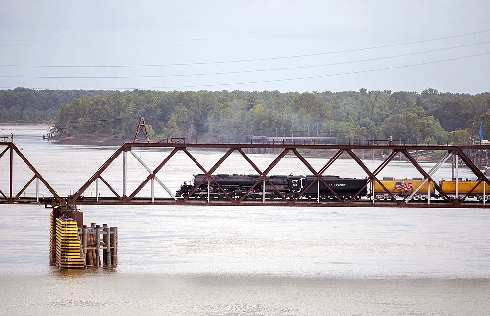 Large steam locomotive on a truss-type steel bridge over a river.