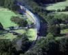 Steam locomotive rounding a curve in a rural area with automobiles and people stopped to watch at a grade crossing.