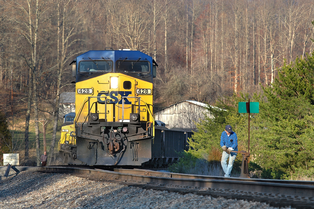 Man throwing switch as train with blue and yellow locomotive waits