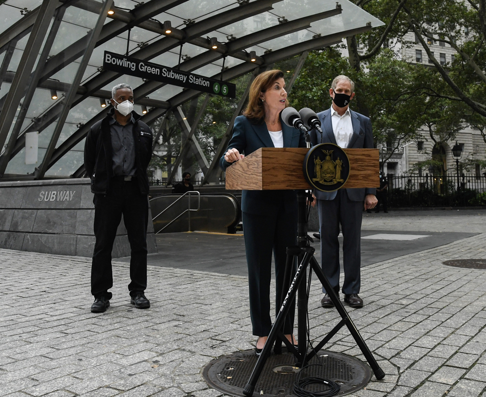 Woman at podium flanked by two men wearing masks in front of subway station