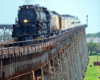 Large steam locomotive with train on long bridge seen with telephoto lens.