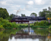 Large steam locomotive rolls over a short deck bridge framed by water, trees, and green shrubs.