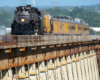 Large steam locomotive with train on a long bridge with a commercial jetliner in the distant background.