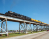 View of a large steam locomotive with yellow passenger train on a bridge, as seen from below.