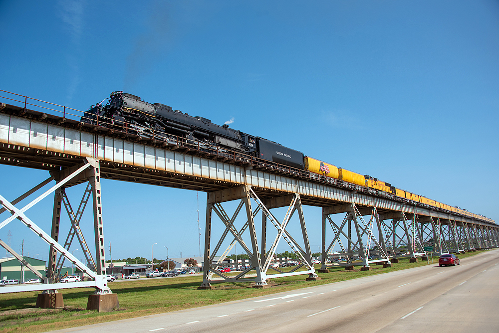 View of a large steam locomotive with yellow passenger train on a bridge, as seen from below.