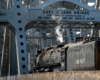 Rear view of a large steam locomotive moving through the trusses of a large steel bridge.