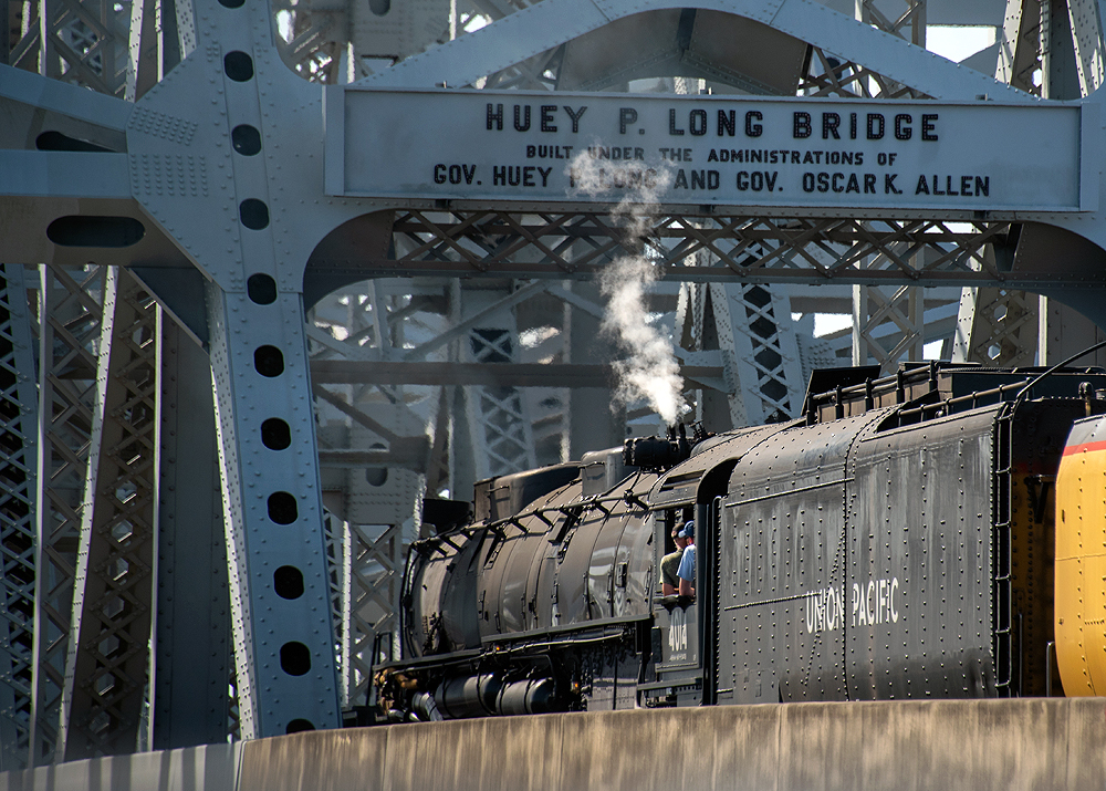 Rear view of a large steam locomotive moving through the trusses of a large steel bridge.