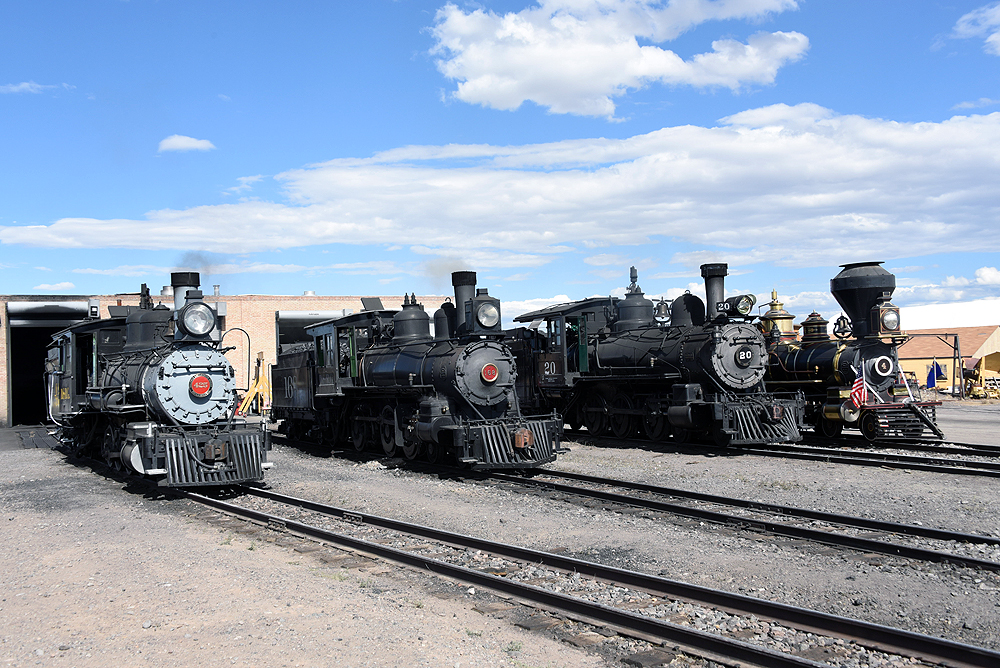 Four steam locomotives side-by-side in a rail yard near an engine house.