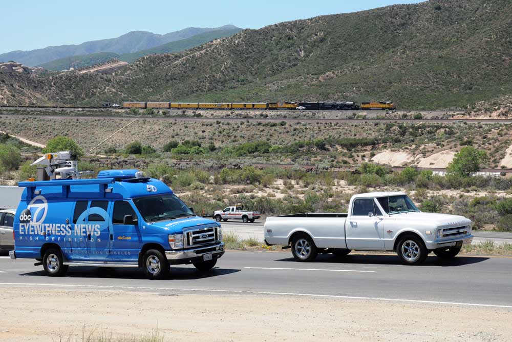 Blue news van and white truck in front of steam locomotive and train