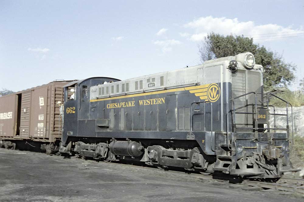 Green and gray diesel locomotive coupled to boxcars with men in white hats in the cab