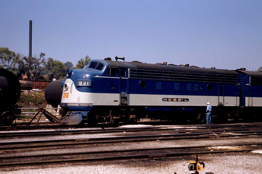 Man in coveralls stands by blue and gray streamlined diesel locomotive in profile