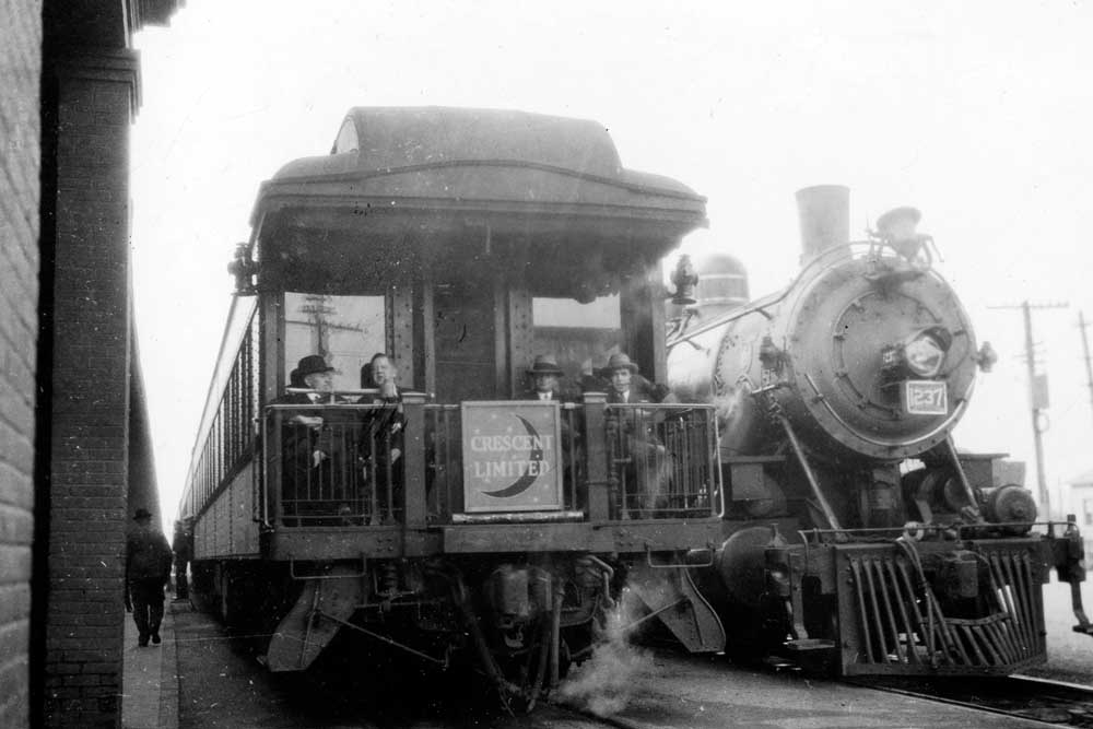 Open platform passenger car aside brick station with steam locomotive nose