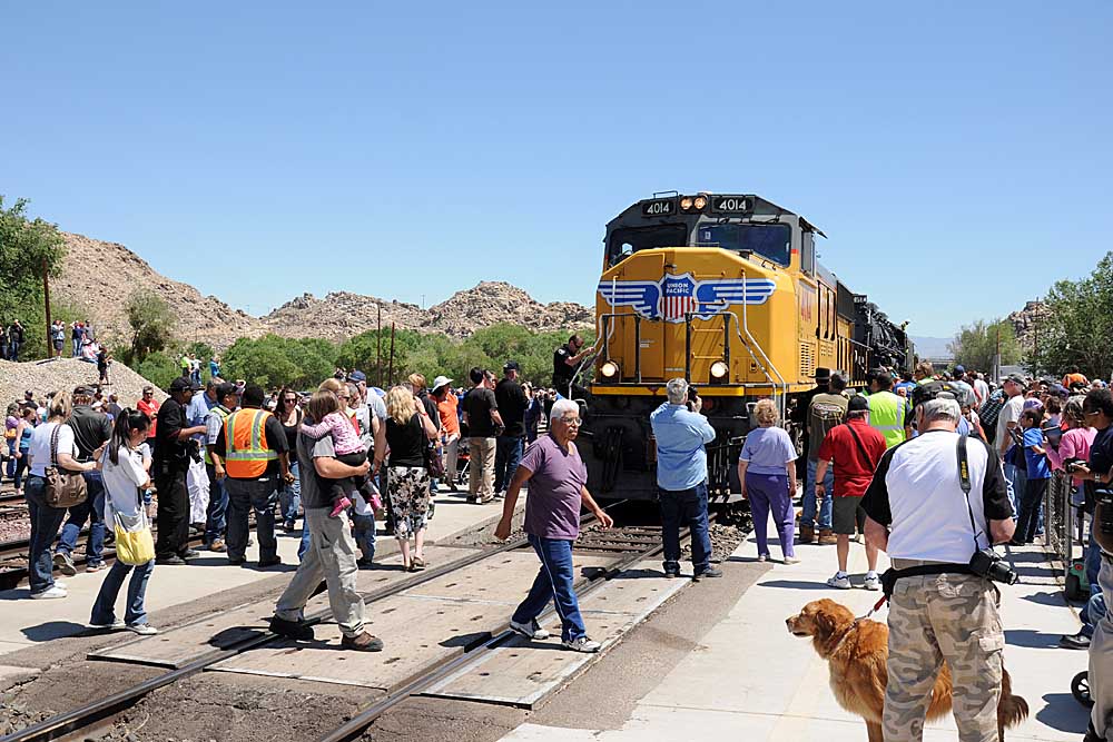 People walk on crossing in front of yellow diesel locomotive