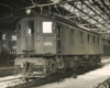 Black-and-white three-quarter-angle photo of box-cab electric locomotive inside shop building.