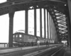 Black-and-white photo of people watching electric locomotives on bridge with freight train